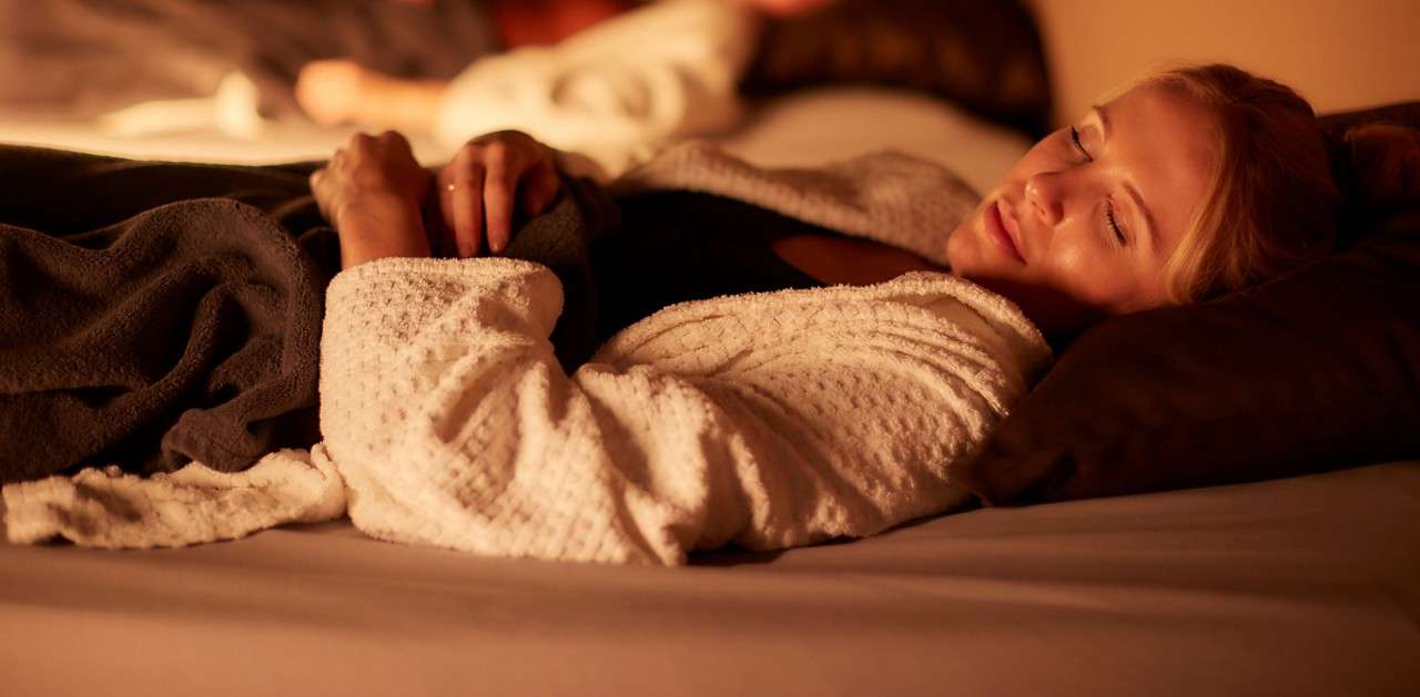 Woman sleeps on a cushioned bed, hands clasping a dark blanket, wearing a white robe; another person rests blurred in background within a warm, dimly lit spa-like relaxation room.