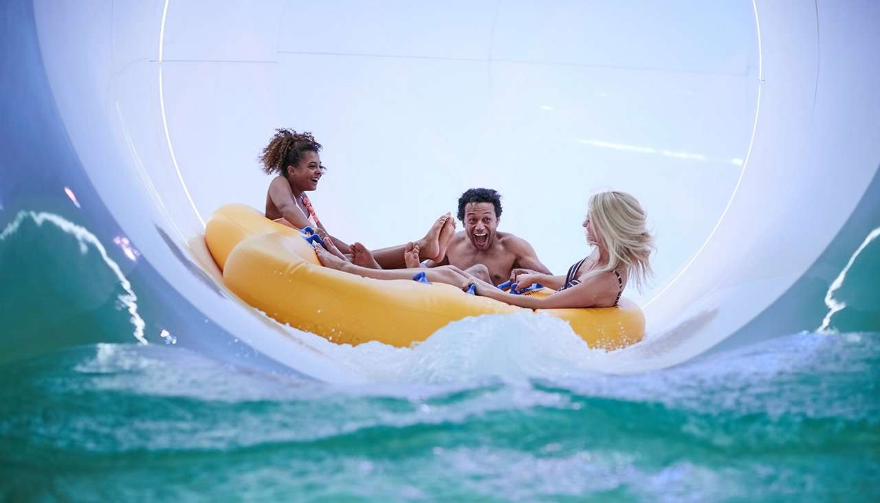 Three riders on a yellow inflatable raft speed and splash, laughing and bracing themselves inside a large enclosed water slide, surrounded by rushing turquoise water and white tunnel walls.
