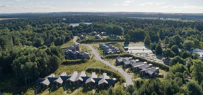 Cabins cluster along winding roads, forming a small resort-like settlement, surrounded by dense forest with parking areas and open courtyards, near distant lakes under a bright daytime sky.