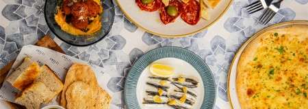Assorted Mediterranean dishes arranged on a patterned tablecloth: cheese pizza, anchovies with lemon, meatballs on puree, roasted tomatoes with olives on toast, and flatbreads, with fork and knife nearby.