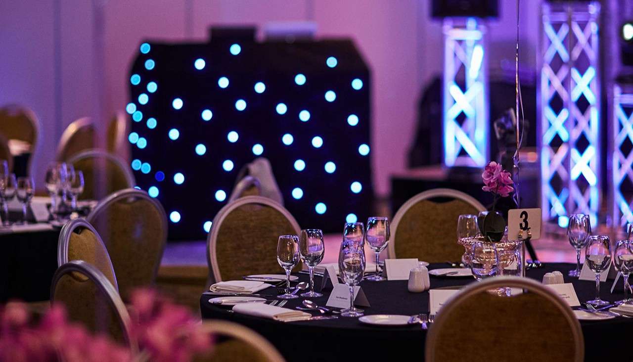 Table set up for event, with black table cloths, pink flowers and sparkling lights in the background