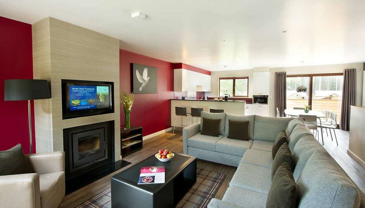 Gray sectional sofa faces a wall-mounted TV above a fireplace, surrounding a low coffee table; beyond, an open-plan kitchen with bar stools and a dining area sits beside large curtained windows.