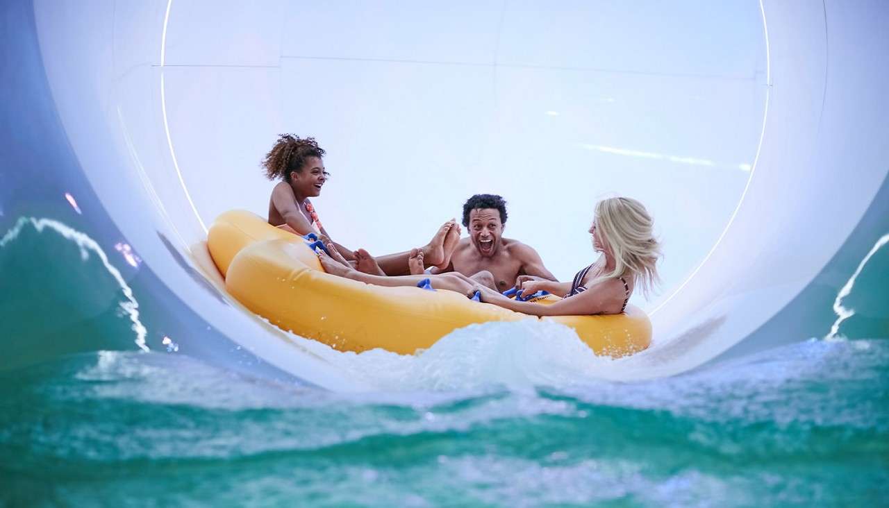 Three people on a yellow inflatable raft ride, laughing and gripping handles, as it rushes through a large enclosed waterslide, splashing into turquoise water in a bright, curved tunnel.