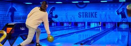 Bowler rolls a green ball down a glowing multi-lane alley toward pins; blue-lit mural surrounds the lanes with silhouettes and large wall text: STRIKE.