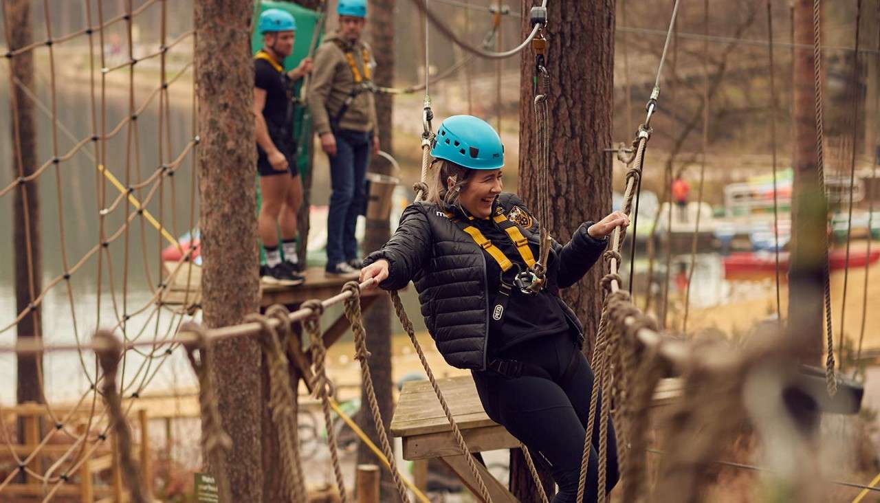 Man balancing across rope bridge between trees as his team mates watch on