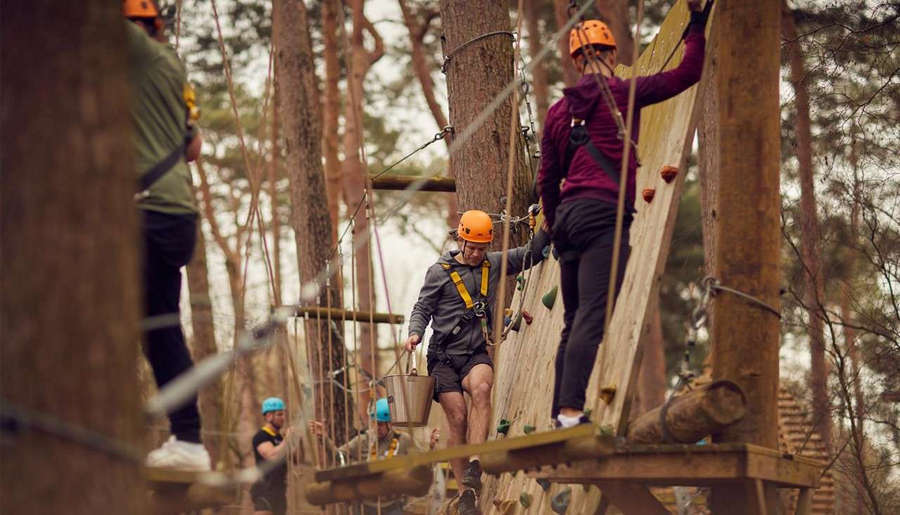 Man carries water-filled bucket across the climbing wall, while his teammate waits to retrieve it at the other side 